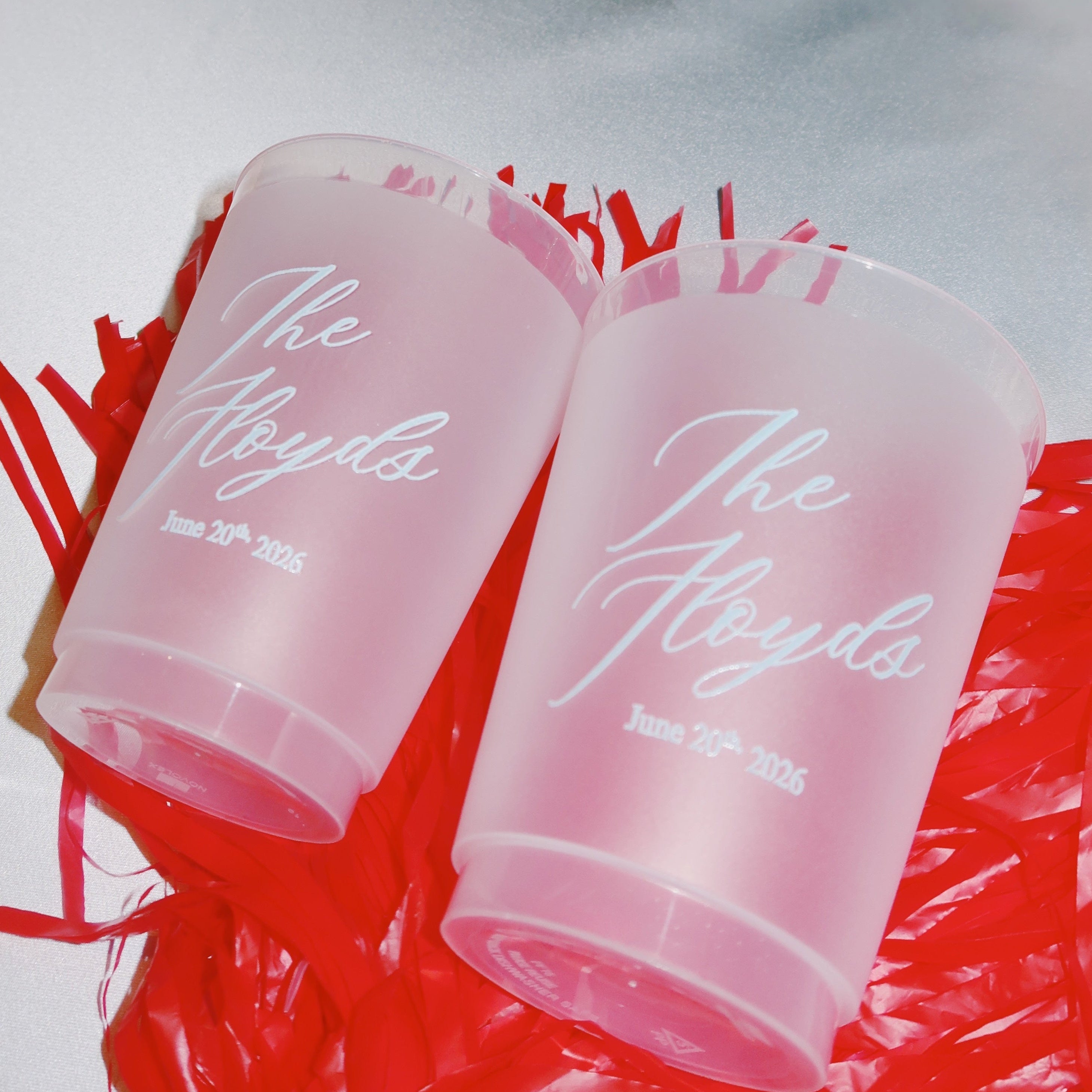 Two personalized frosted wedding cups featuring the couple’s names and wedding date, displayed on a red pom pom background.
