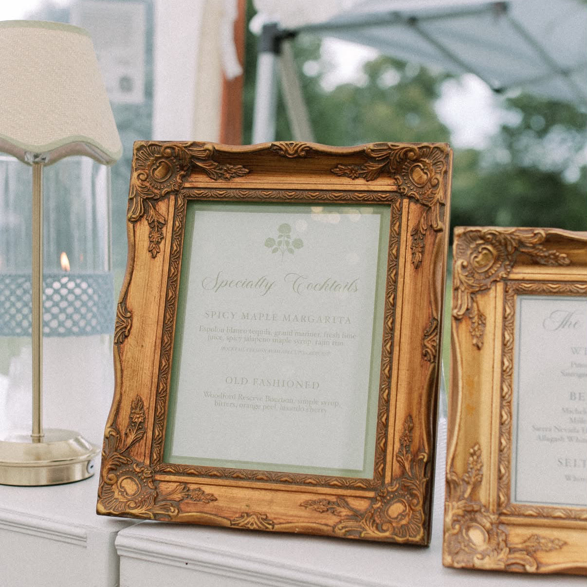 Two ornate gold picture frames displaying bar menu and specialty cocktail menu cards, styled on a white table next to a lamp in an outdoor event setting.
