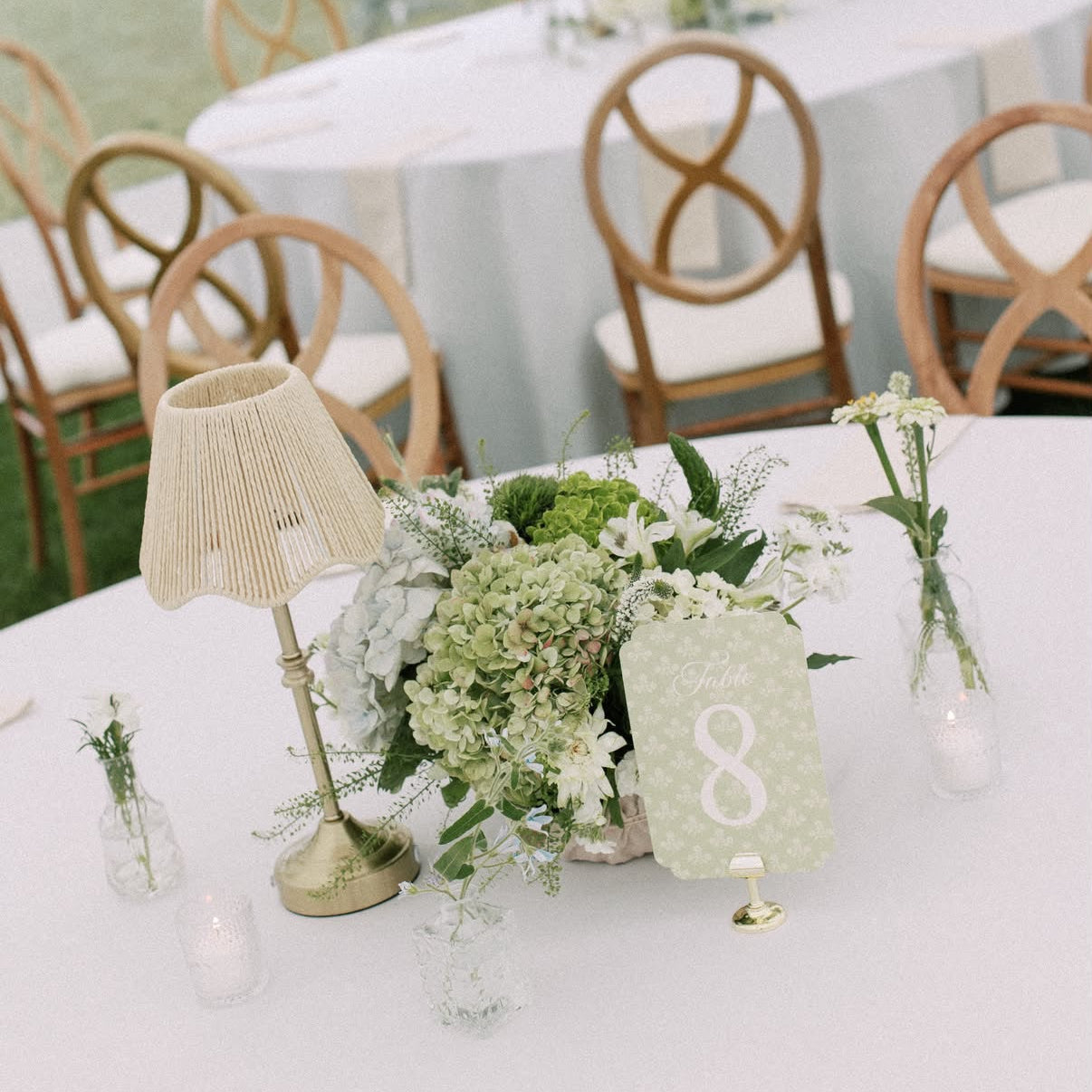 A styled wedding table centerpiece with a cream pleated table lamp, a small floral arrangement in a glass vase, a green table number card showing the number 8, and clear glass candle holders on a white tablecloth. In the background, other decorated tables and wooden chairs are visible inside a tent outdoors.