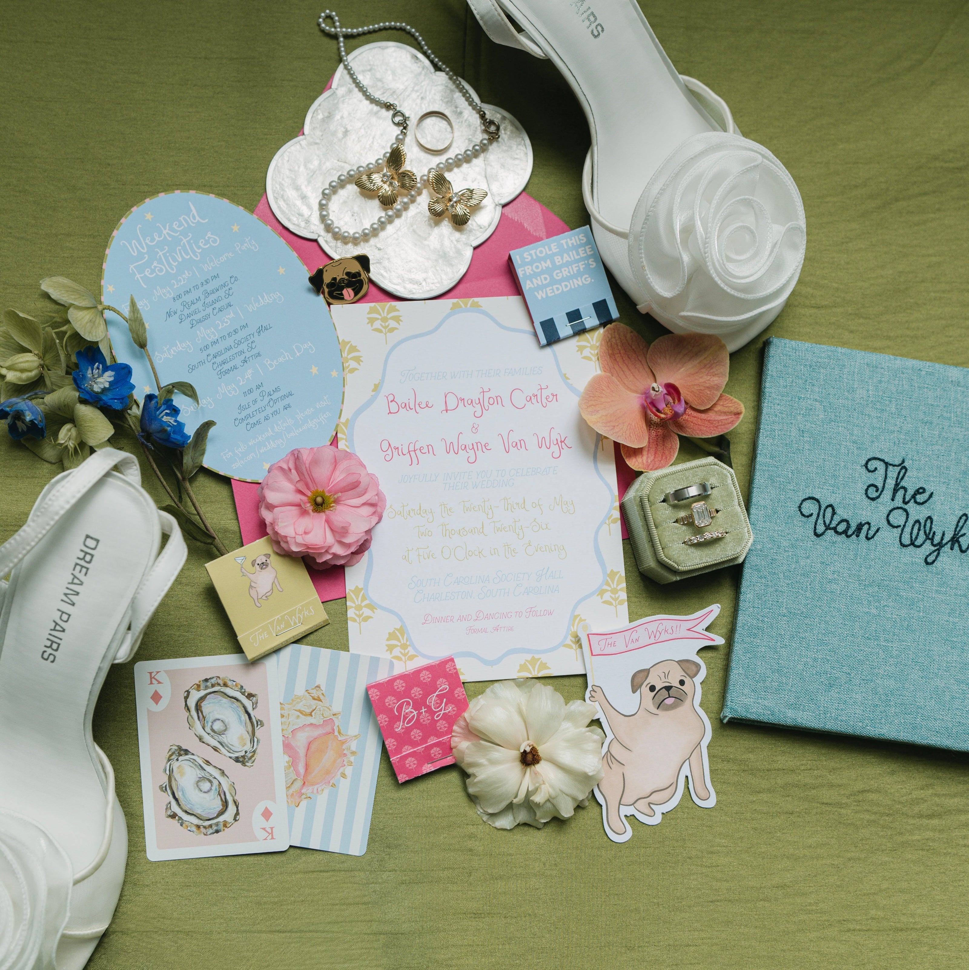 Flat lay featuring a custom wedding invitation suite with floral details, bridal shoes, embroidered keepsake box, personalized matchbooks, and wedding day accessories.